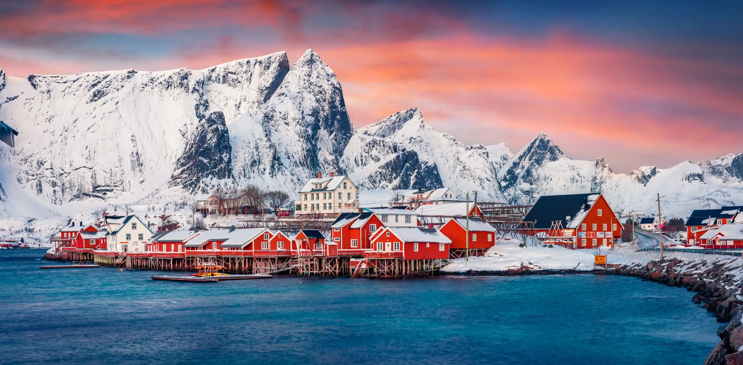 Rote Rorbuer-Hütten am Wasser vor schneebedeckten Lofoten-Bergen im rosafarbenen Winterlicht